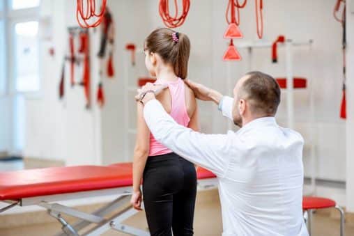 chiropractor looking on the back of the junior girl during the medical examination at the rehabilitation office with suspension medical equipment